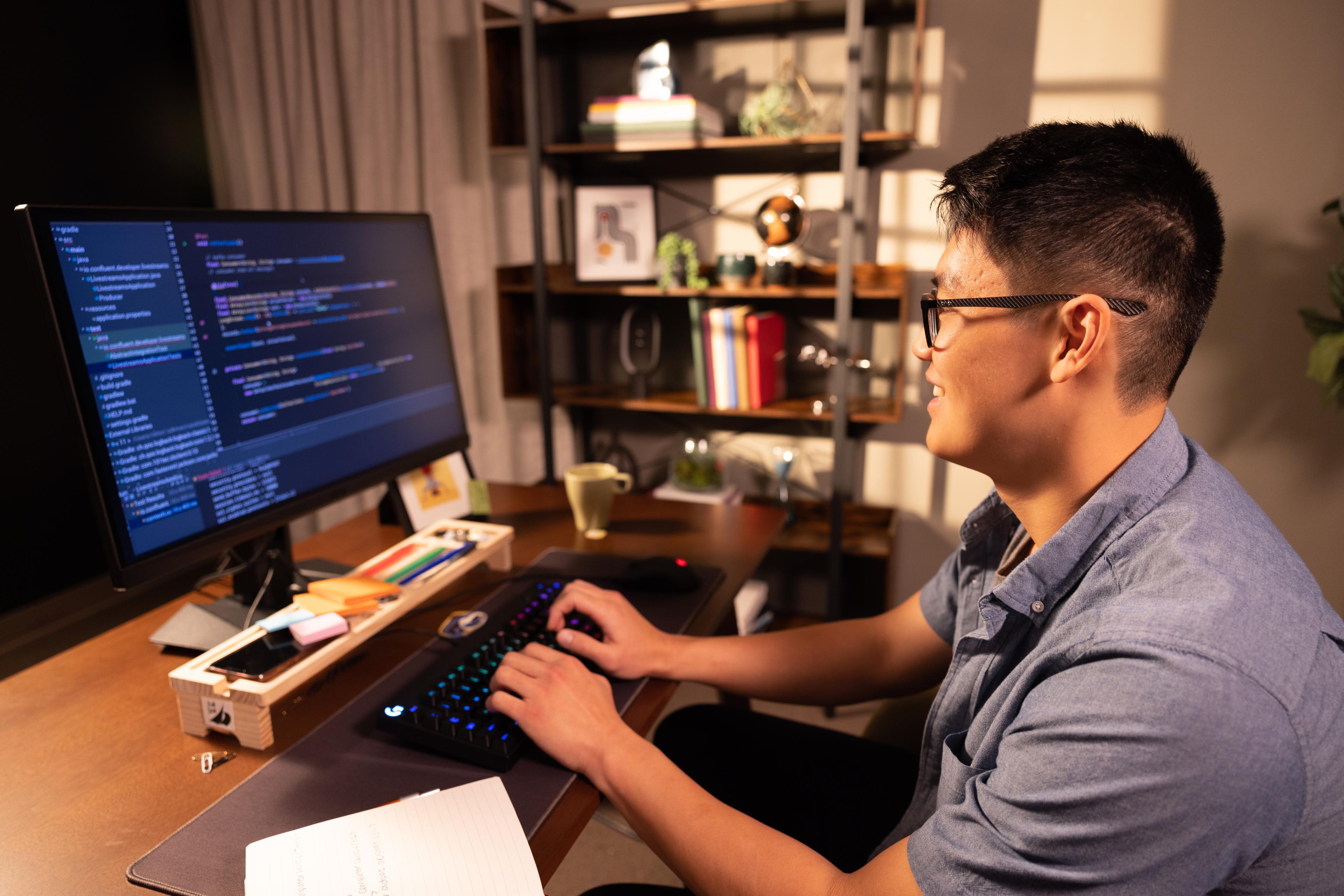 A smiling student sits at a desktop computer in a home office and types lines of code.
