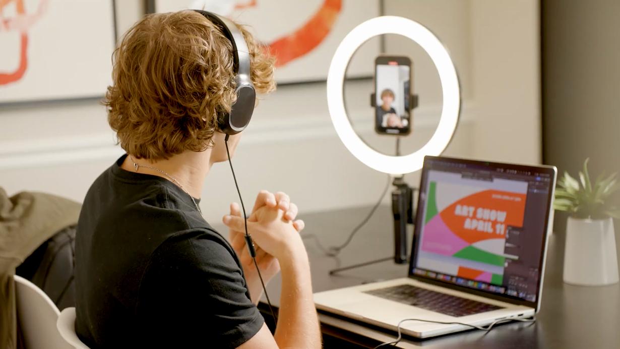 A student sits at a desk and uses a laptop, smartphone, and ring light to film video content.
