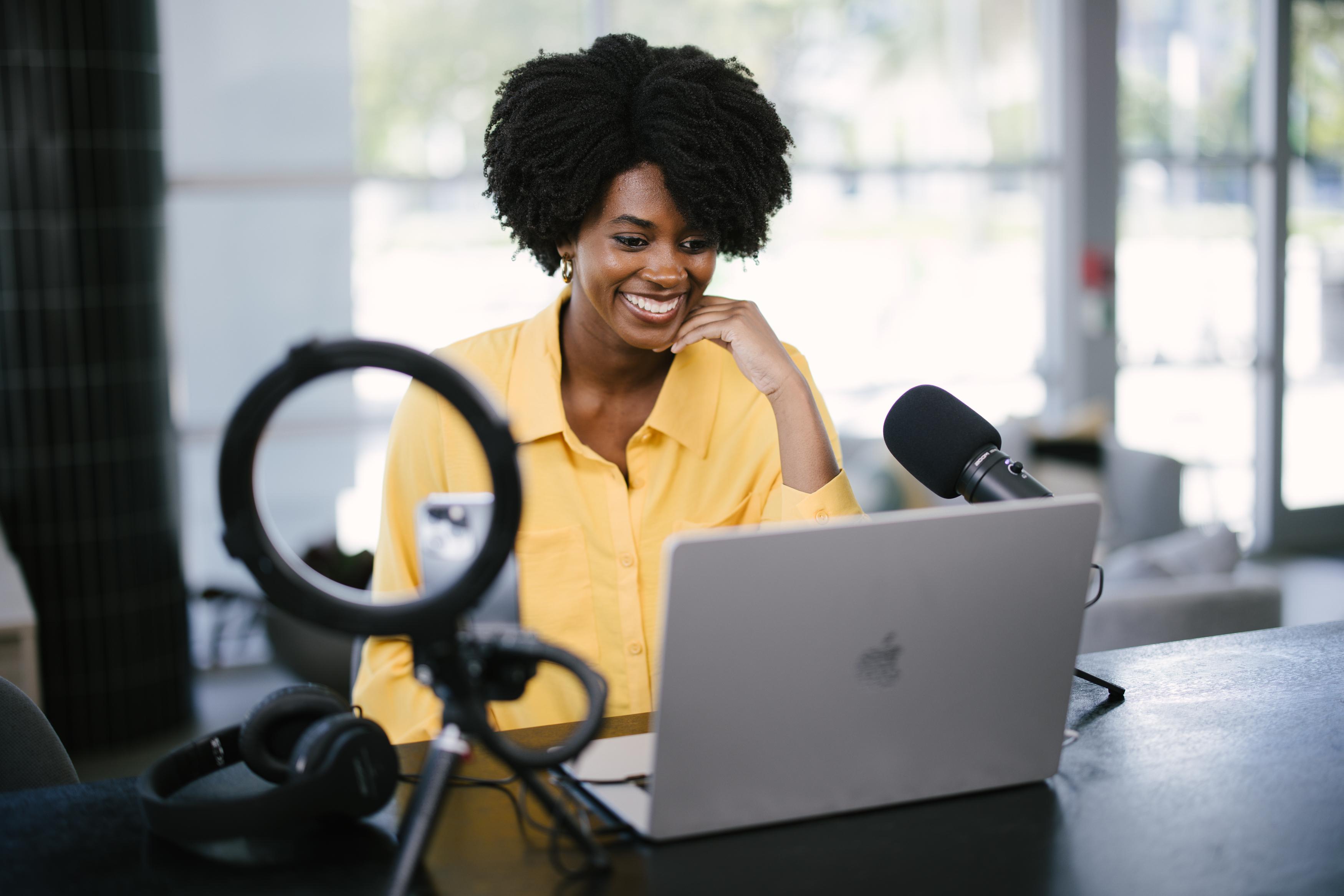 A smiling student in a bright yellow shirt sits at a desk with a laptop, microphone, and ring light.