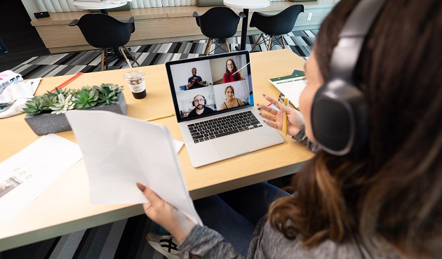 A student wearing headphones sits at a table and attends a Zoom meeting on their laptop.