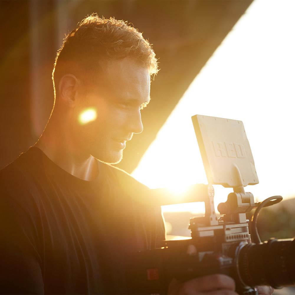 A man in a dark shirt stares into the screen of a video camera, while the sun sets behind him.