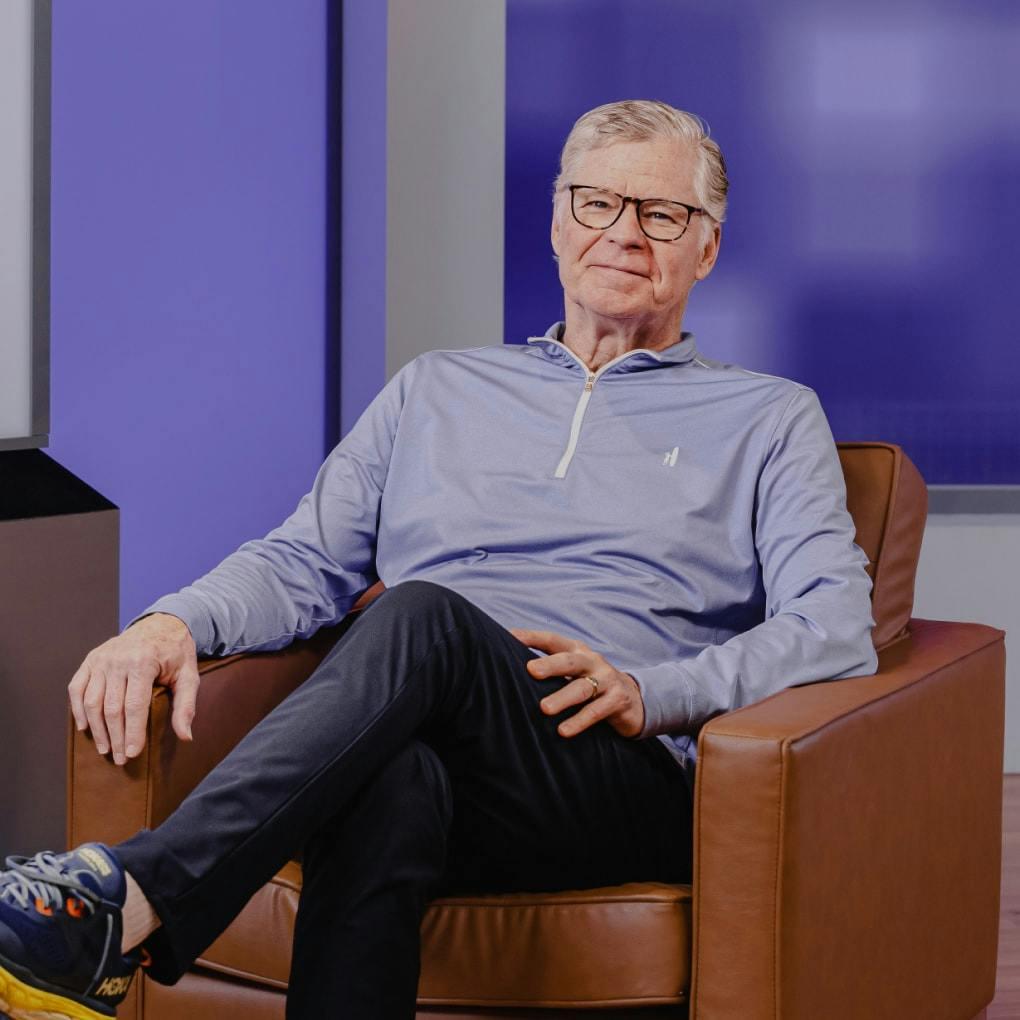 Dan Patrick sits in a brown leather armchair. He is smiling and wearing a light blue athletic sweater, dark pants, and sneakers. There is a television behind him that reads: Full Sail DC3.