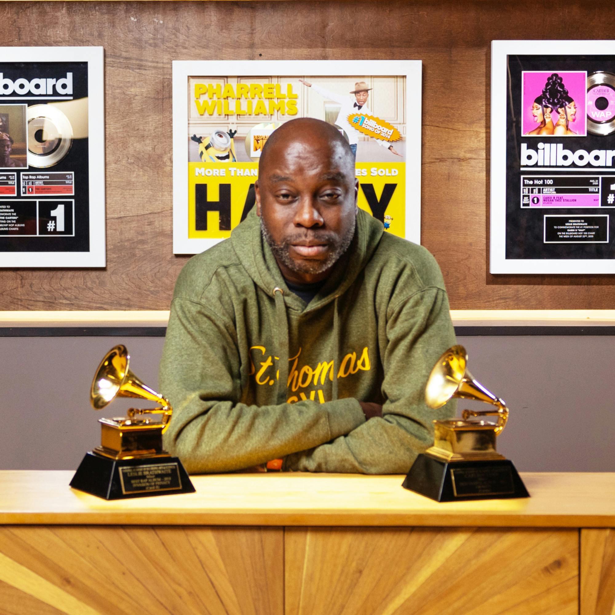 Leslie Brathwaite, wearing a green hoodie, sits with his arms folded on a desk with two of his Grammy awards in front of him. Behind him are framed gold records and billboard awards of songs he has mixed.